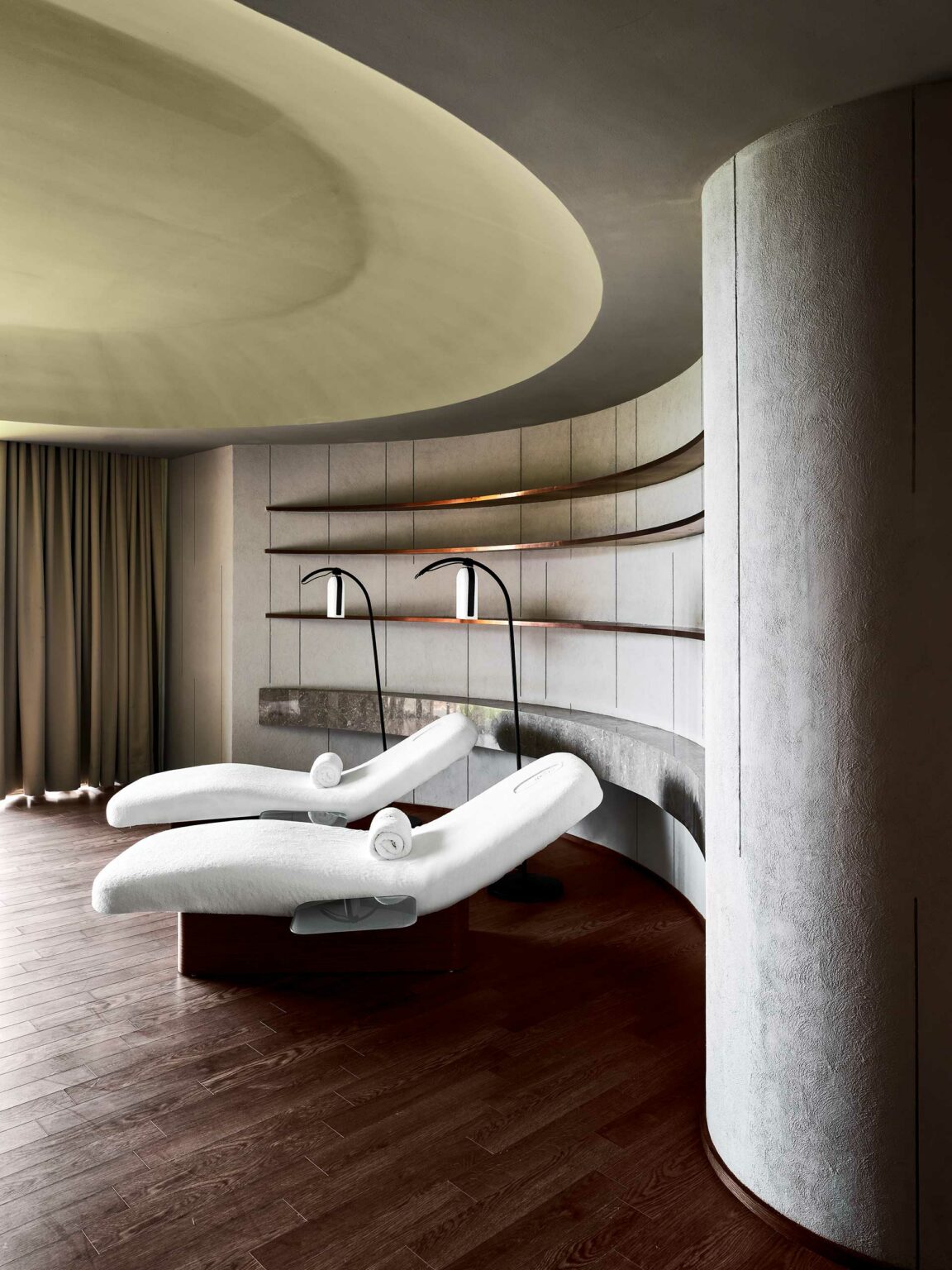 Modern spa room with two white chaise lounges and rolled towels on dark wooden floors. Curved walls feature minimalist shelves and lamps, creating a serene ambiance.