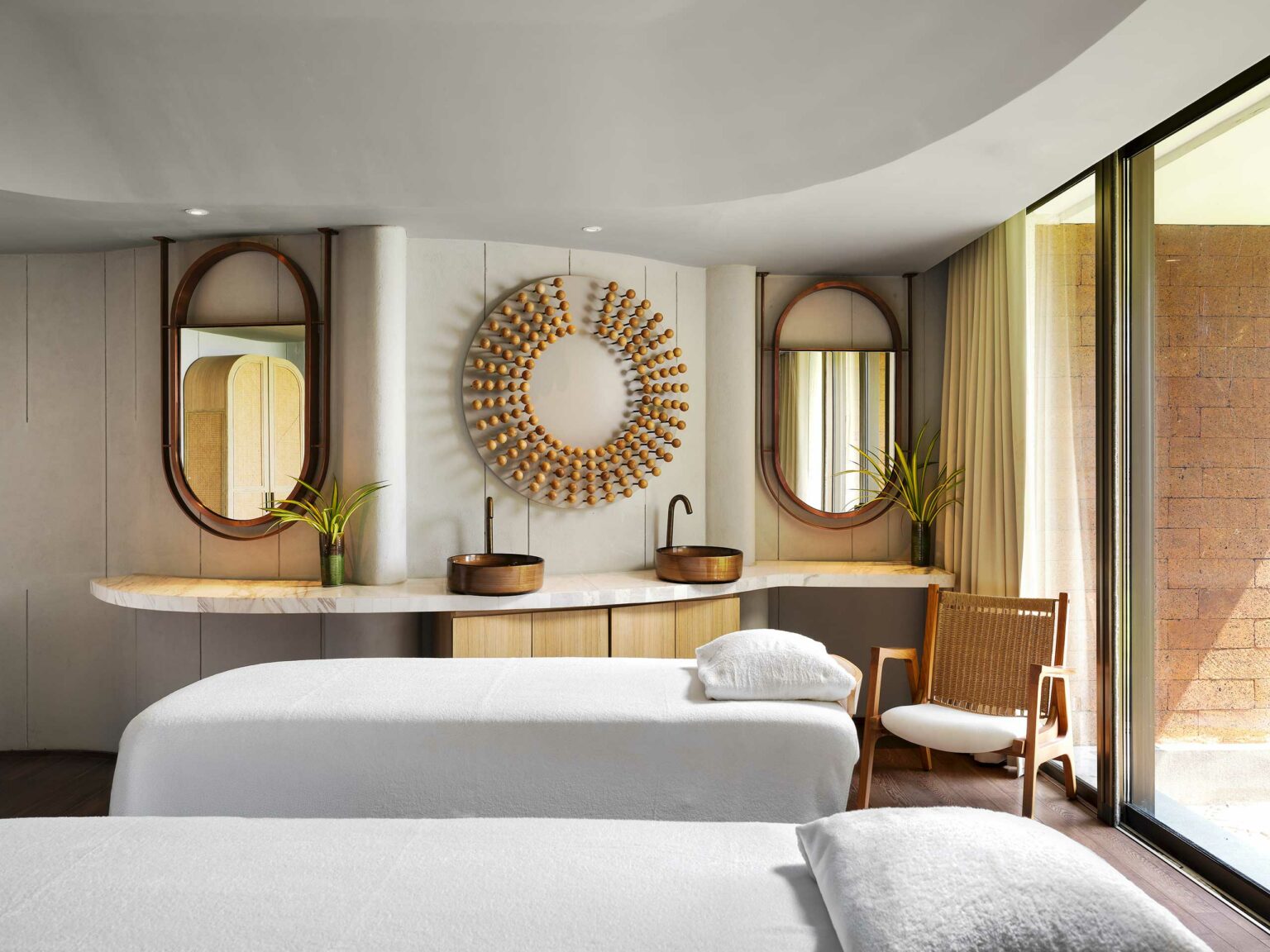 A serene spa room with two treatment beds, elegant decor, oval mirrors, and a sunburst wall art. Warm lighting enhances the calm atmosphere.