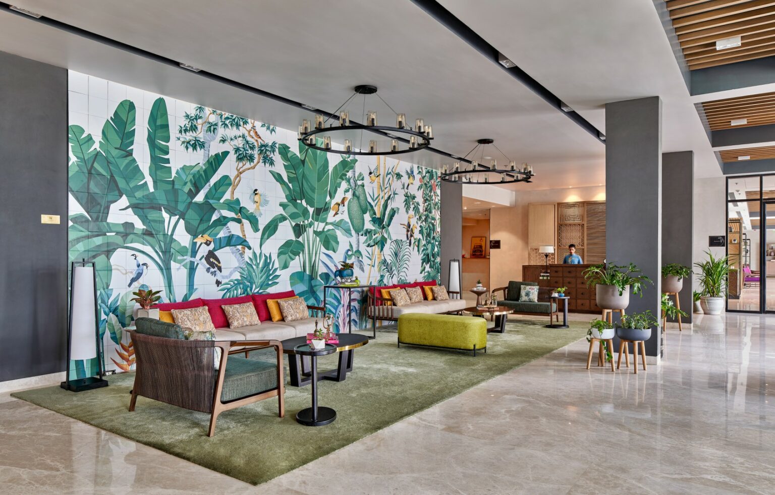 Modern hotel lobby with vibrant tropical mural of large green leaves. Features plush seating with colorful cushions, sleek lighting, and a welcoming atmosphere.