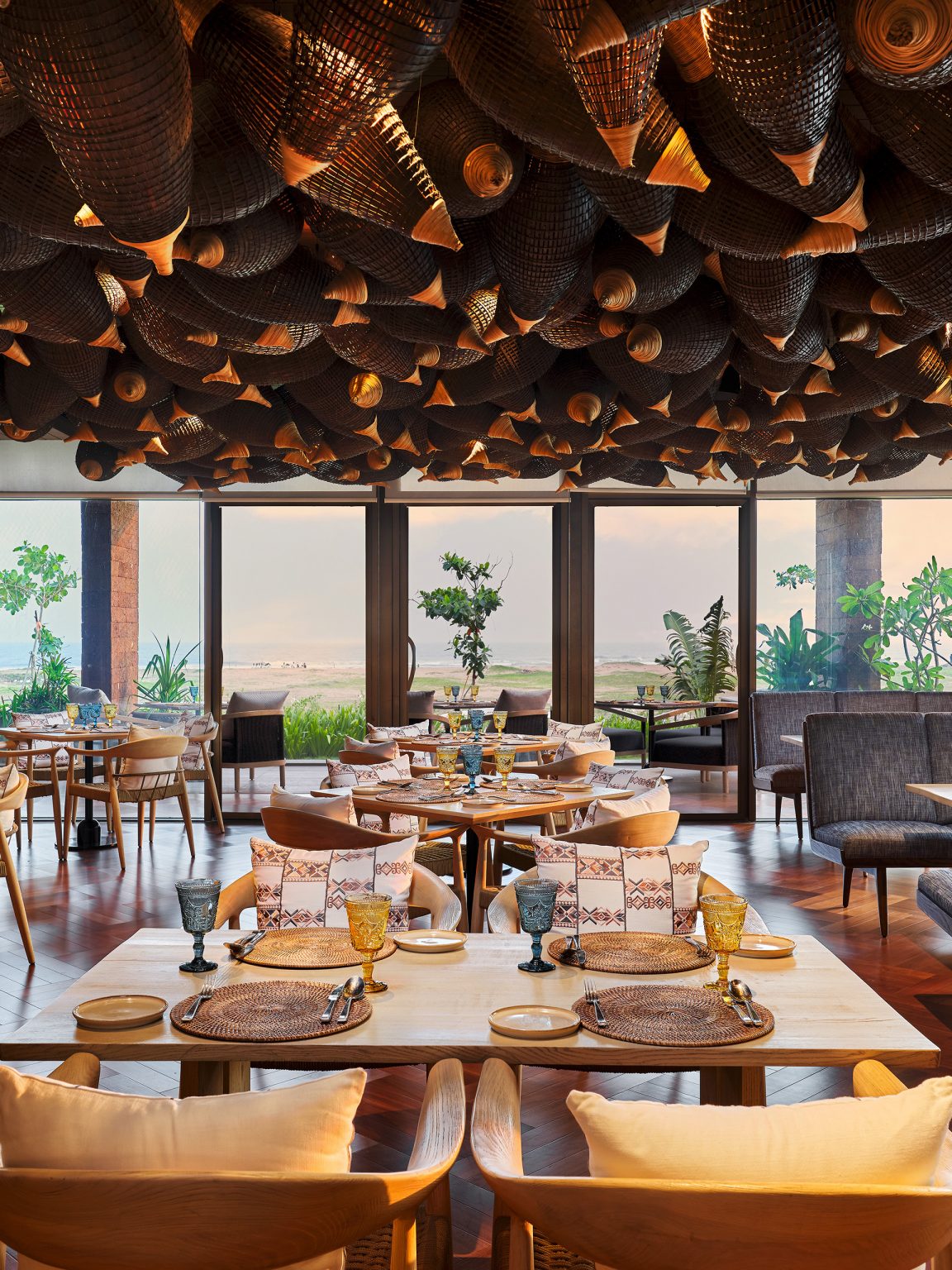 Elegant restaurant interior with a warm, sophisticated vibe. Features woven decor on the ceiling, wooden floors, modern tables, cushioned chairs, and large windows with a scenic view.