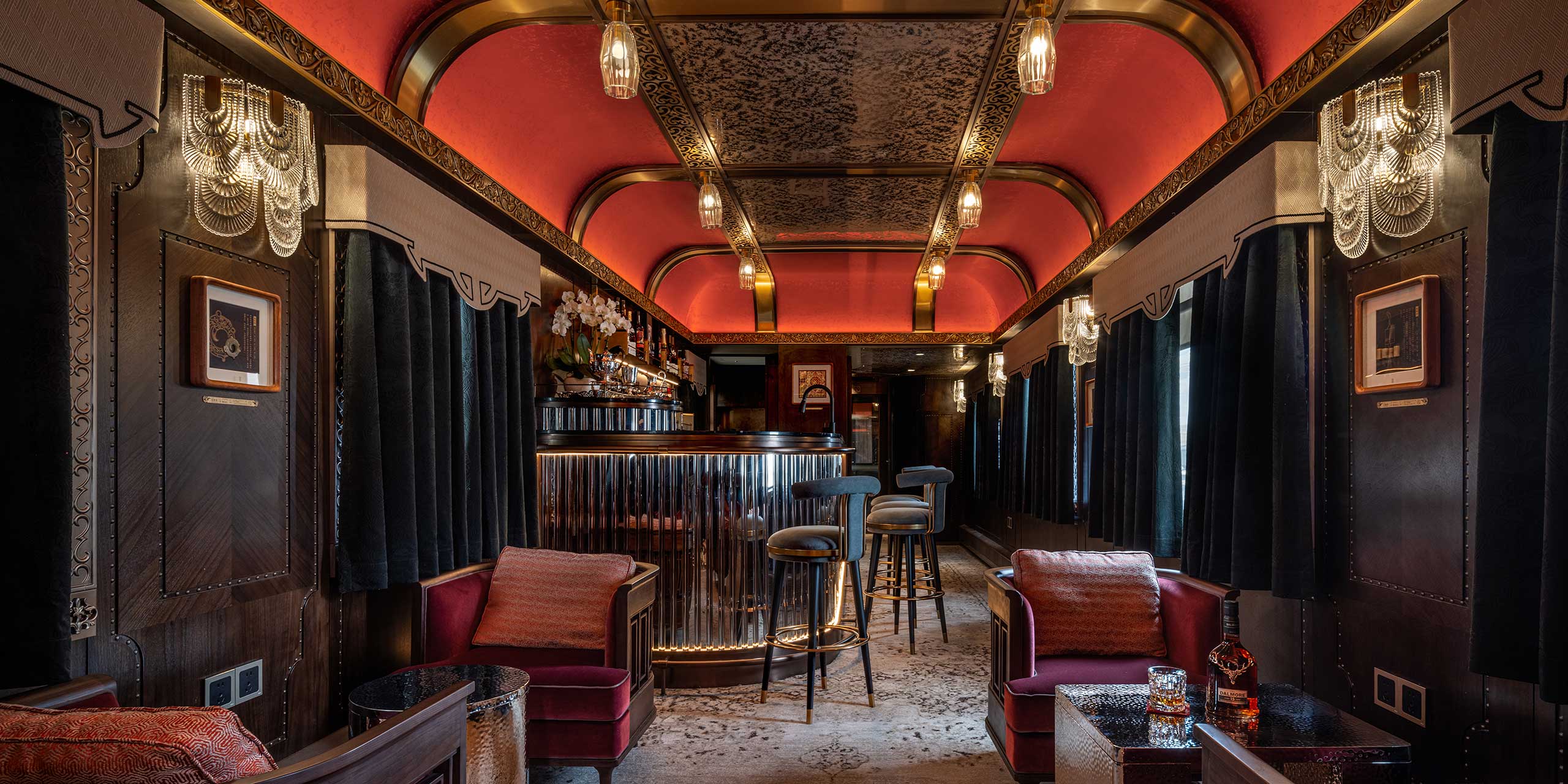 Luxurious train car bar with red ceilings, ornate chandeliers, and a gleaming counter. Plush seating and a warm, elegant ambiance create a vintage feel.