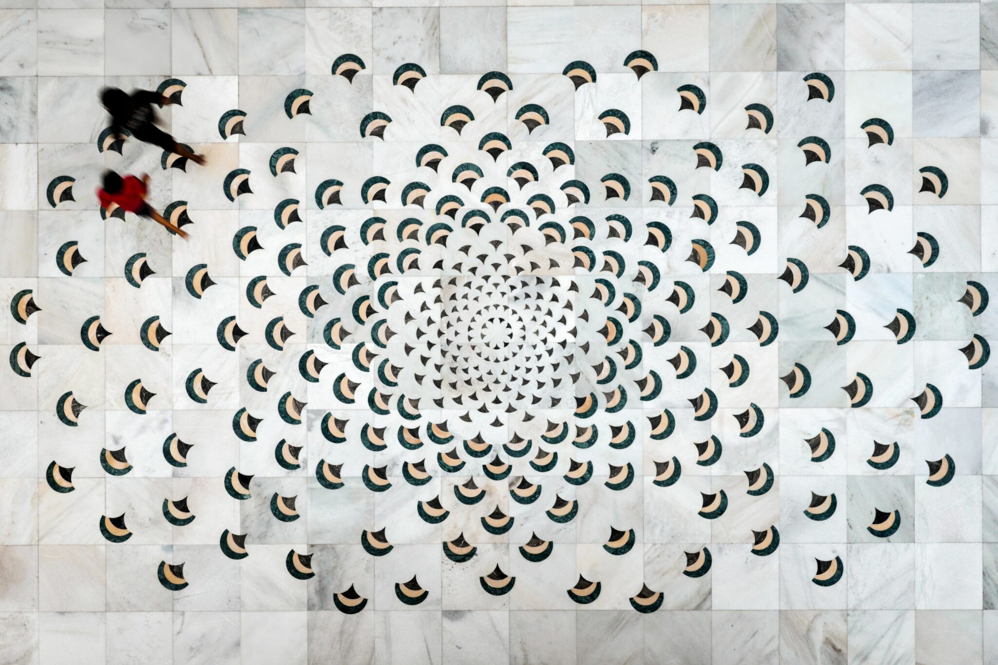 Aerial view of a geometric, spiral pattern on a marble floor with repeated fan shapes. Two blurred figures walk across, adding dynamic motion.
