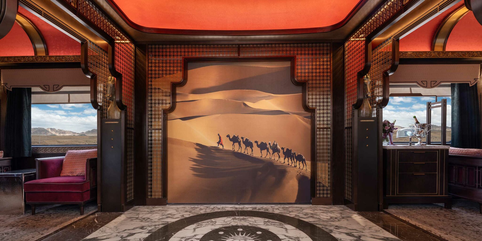 Luxurious train interior with ornate decor. Central mural depicts camels crossing a desert. Rich red tones, plush seating, and elegant details create a lavish atmosphere.