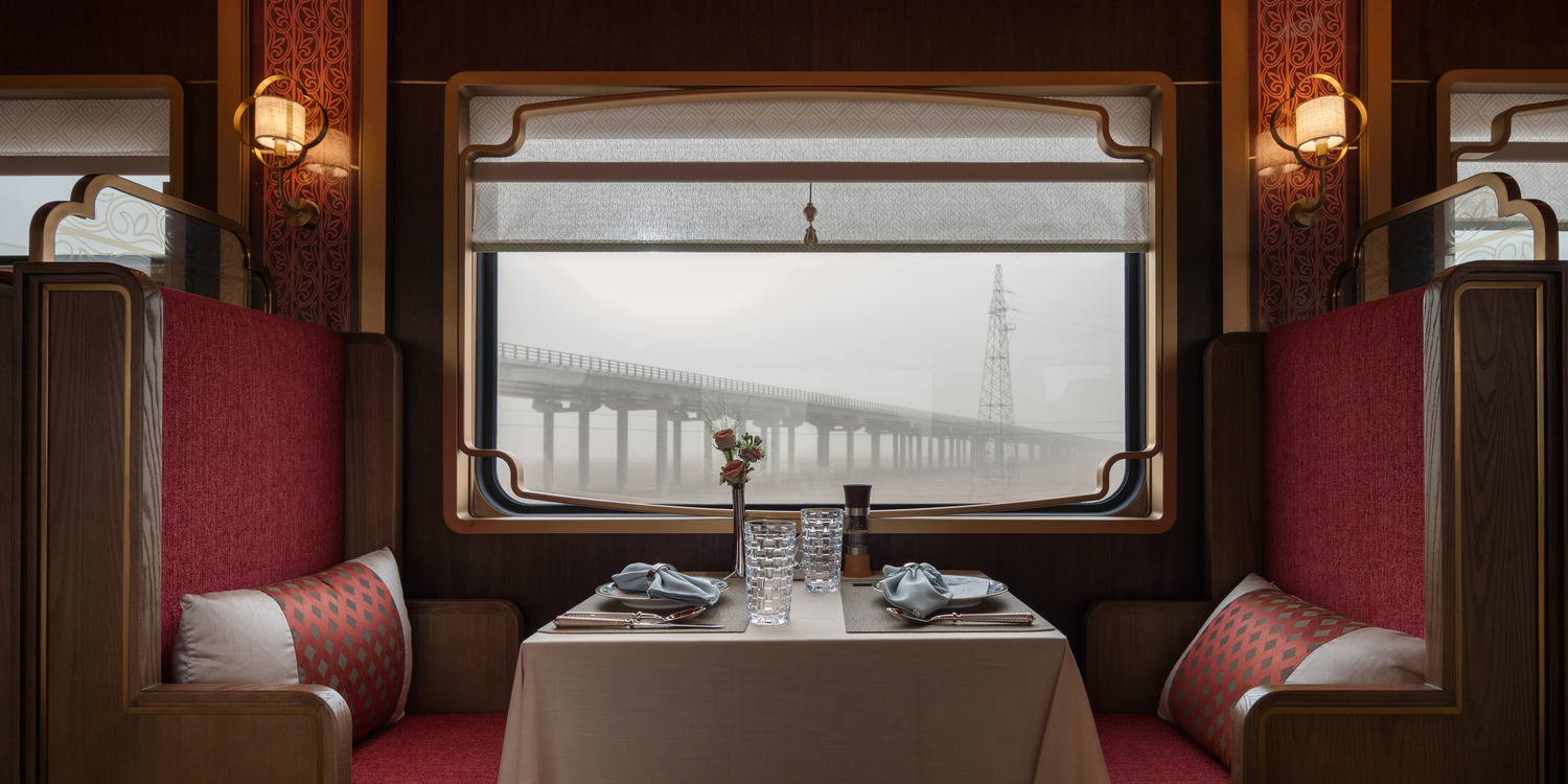 Elegant train dining booth with red seats and floral cushions. A table set for two with crystal glasses and a flower in a vase. Misty bridge scene outside.