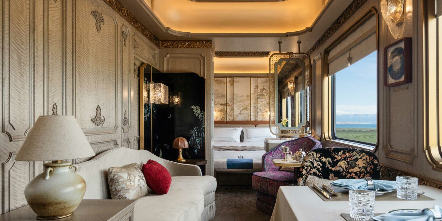 Luxurious train cabin with ornate wood paneling, a plush sofa, and dining table. Large window reveals a serene landscape, creating a cozy, elegant atmosphere.
