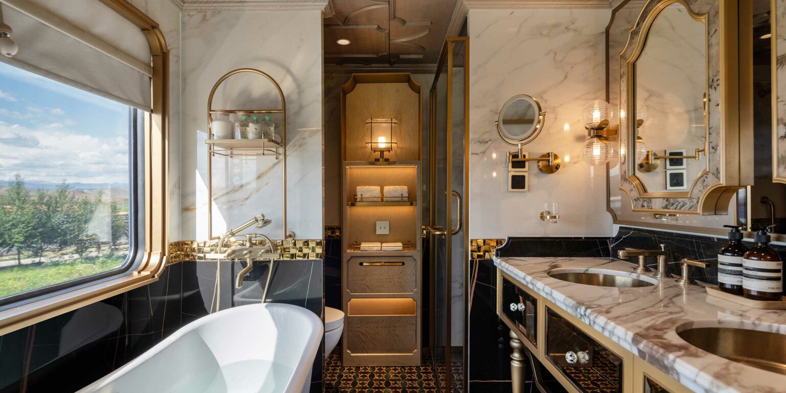 Luxurious train bathroom with marble and gold accents, featuring a freestanding tub, dual sinks, large mirrors, and an elegant window view.