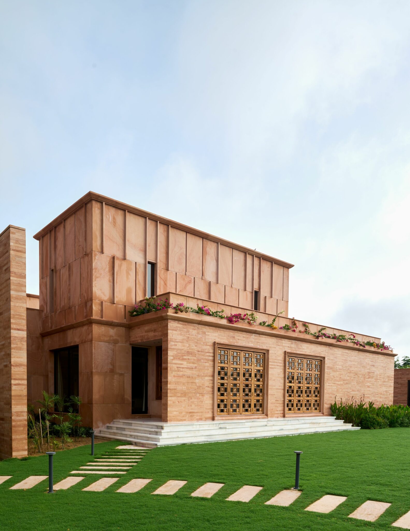 A modern two-story house with a warm sandstone facade and decorative wooden panels. It features a lush green lawn with stepping stones, and a vibrant rooftop garden. The atmosphere is serene and elegant.