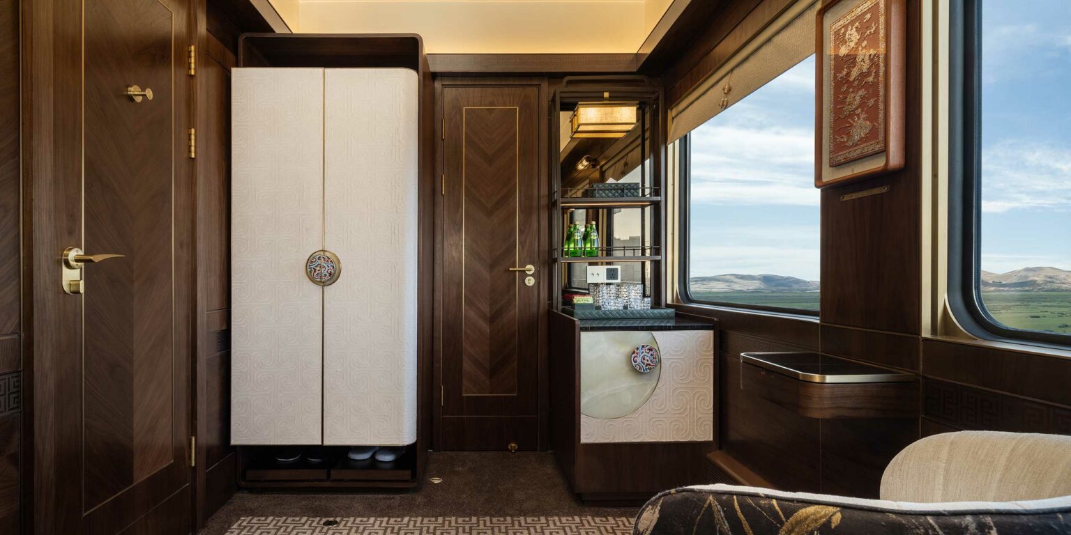Luxurious train cabin interior with elegant wood paneling and warm lighting. Features a minibar with green bottles, plush seating, and scenic window view.