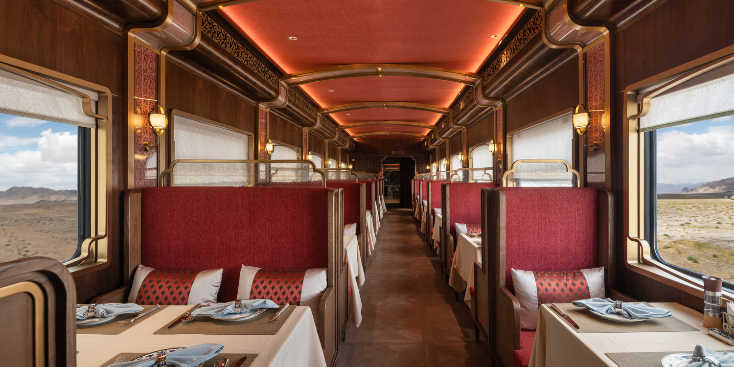 Luxurious train dining car with plush red booths and elegant table settings. Large windows reveal a scenic desert landscape. Warm, inviting ambiance.