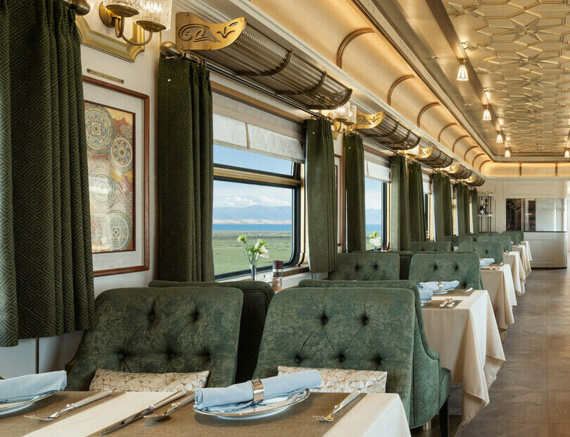 Luxurious train dining car with elegant green chairs, white tablecloths, and large windows revealing scenic countryside views. The ambiance is classic and re