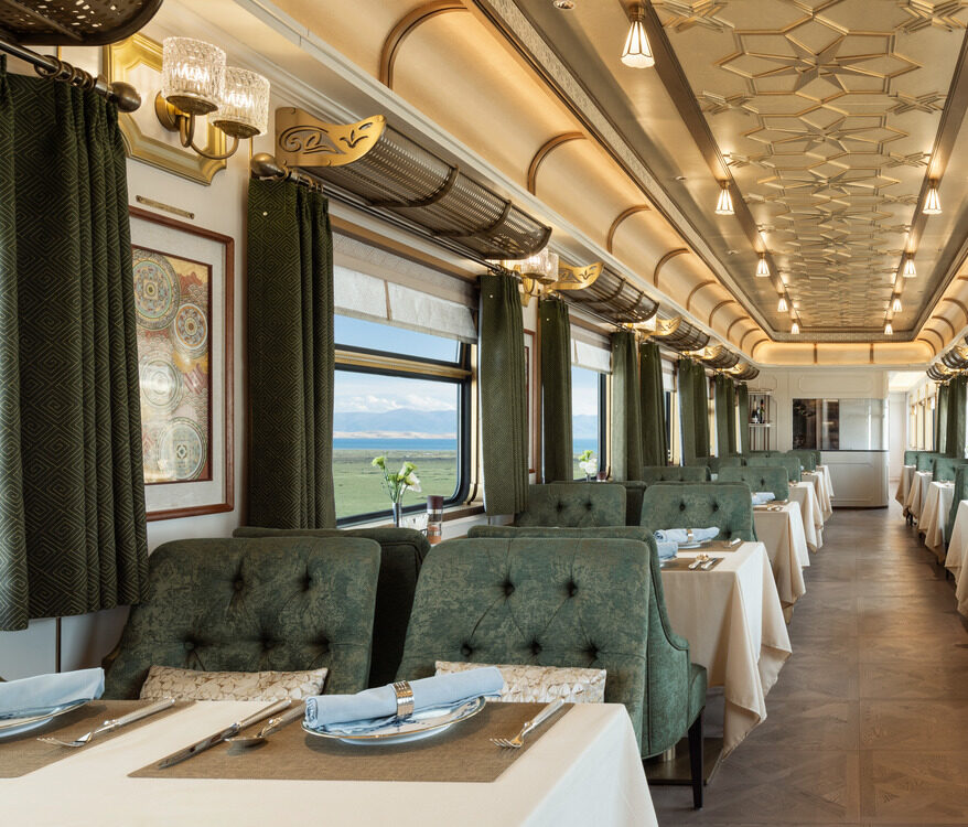 Luxurious train dining car with elegant green chairs, white tablecloths, and large windows revealing scenic countryside views. The ambiance is classic and re