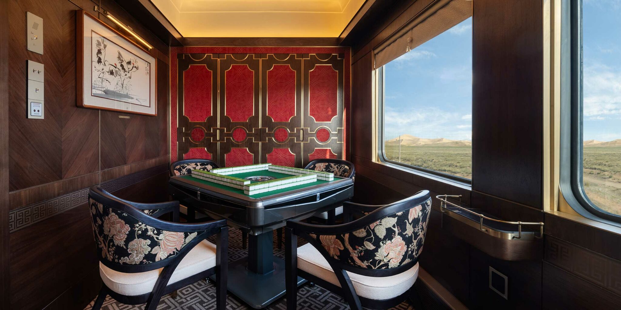 Luxurious train interior featuring a games table with floral chairs, ornate red and gold paneling, and a landscape view through a large window.