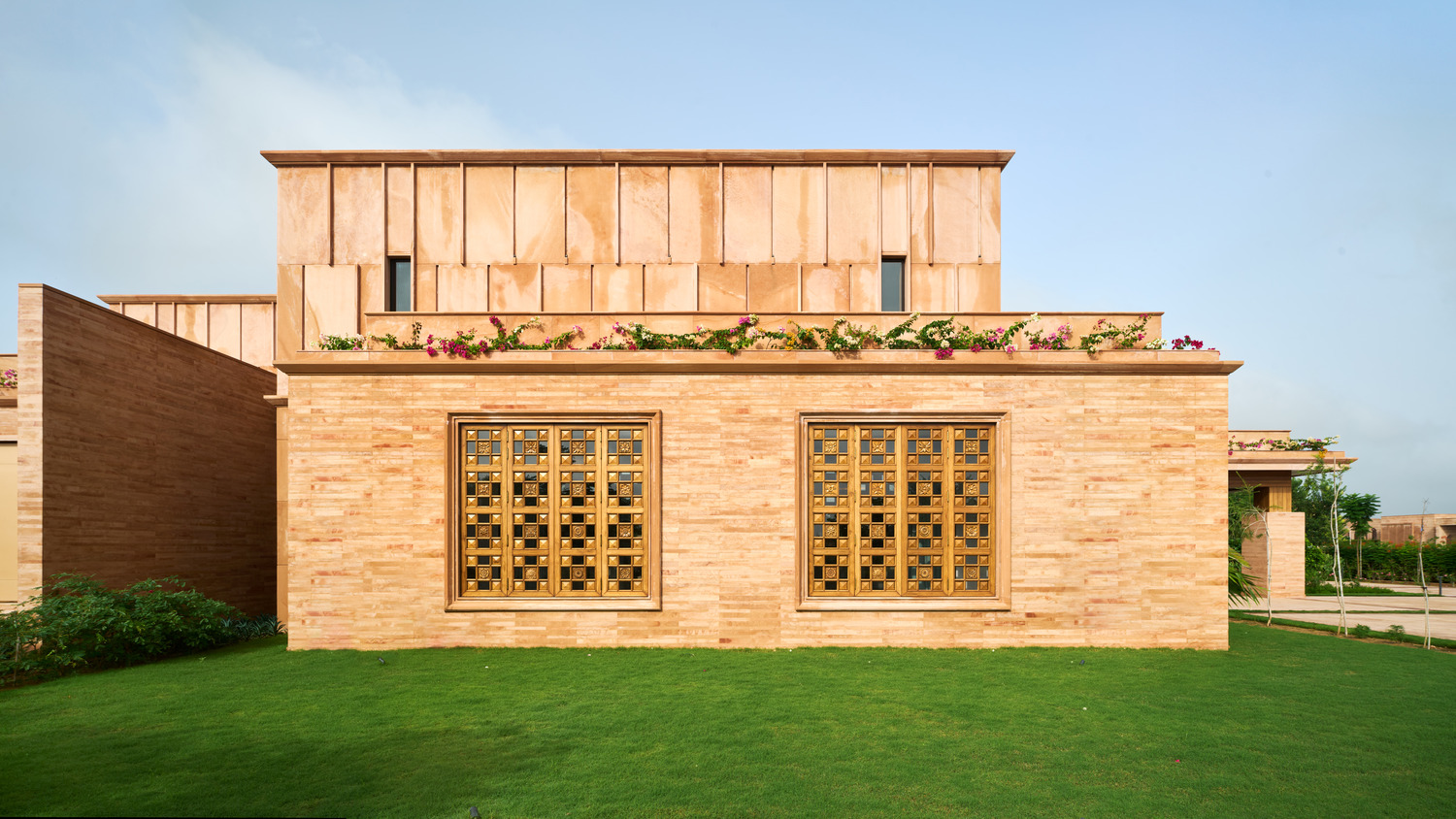 A modern beige stone building with two large, ornate windows, surrounded by lush green grass and flowered plants on top, under a clear blue sky.