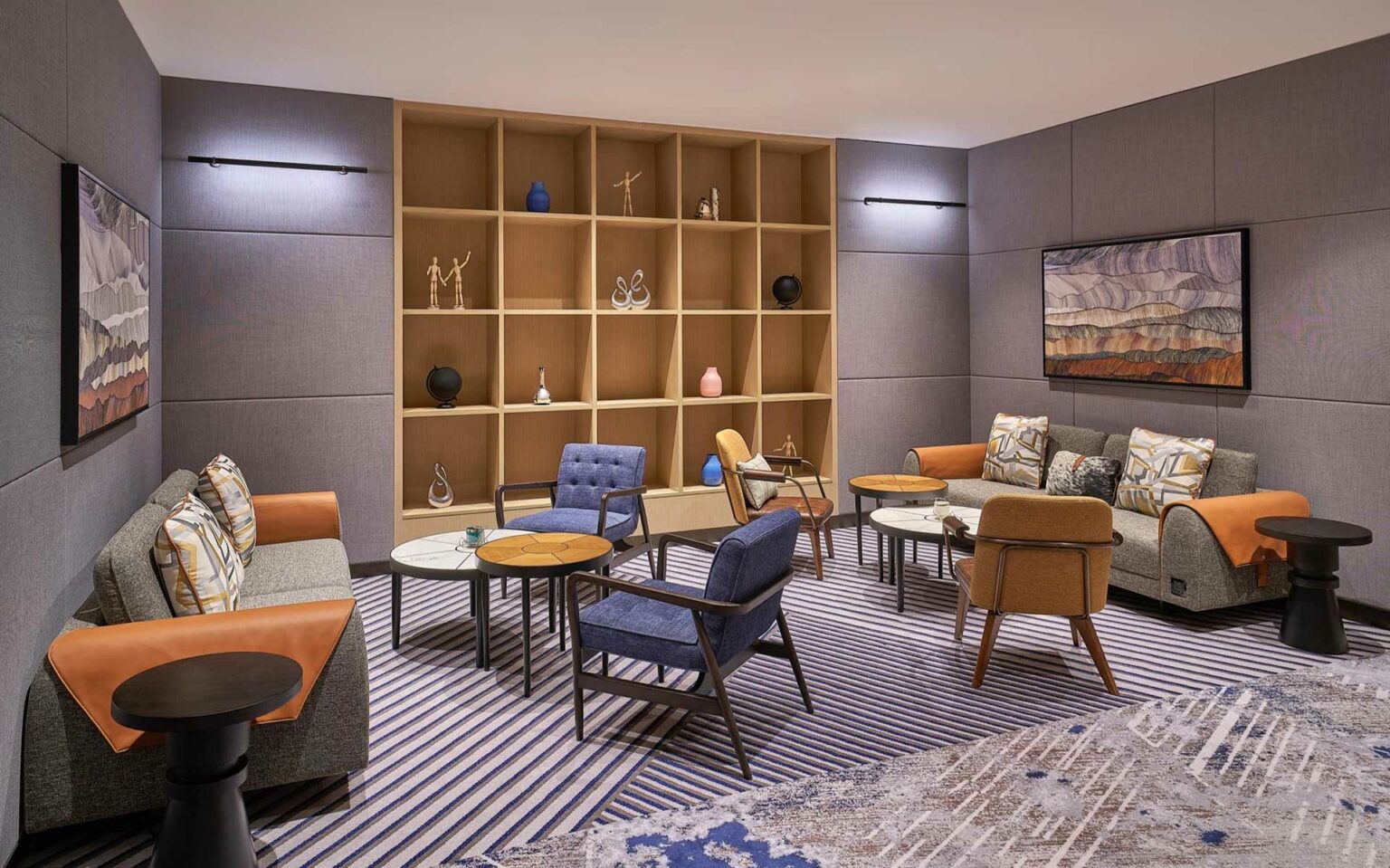 Modern lounge with gray walls features a wooden bookshelf with decor items. Cozy seating includes patterned sofas and blue chairs. Contemporary art adorns the walls.