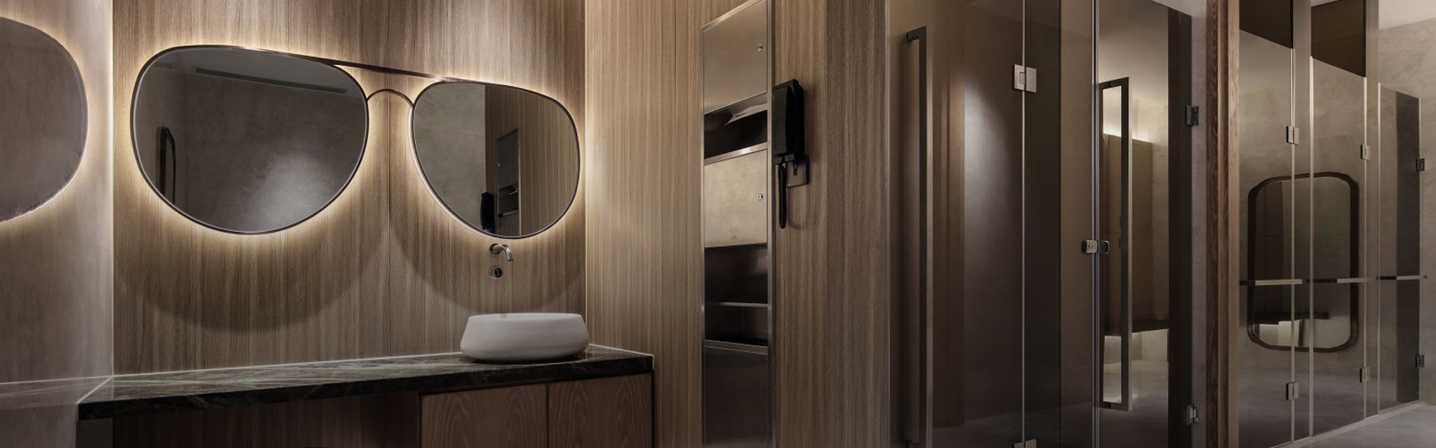 Modern bathroom with wood paneling and sleek glass doors. A large mirror shaped like sunglasses is above a dark countertop and white basin, creating a stylish and unique atmosphere.