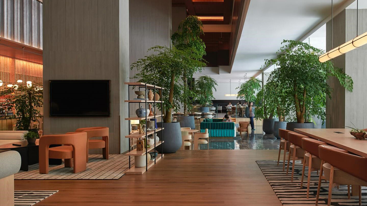 Modern hotel lobby with wooden floors, large potted plants, orange chairs, and a teal couch. A warm, inviting atmosphere with ample natural light.