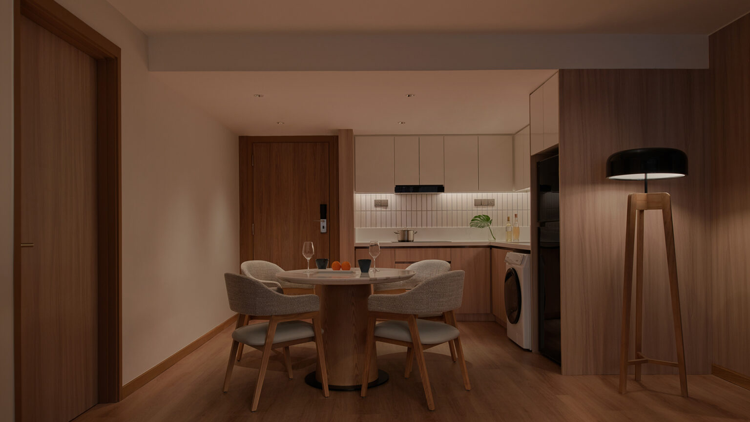 A cozy, dimly lit dining area features a round wooden table with four upholstered chairs. A modern kitchen is in the background. Warm lighting creates a relaxed atmosphere.