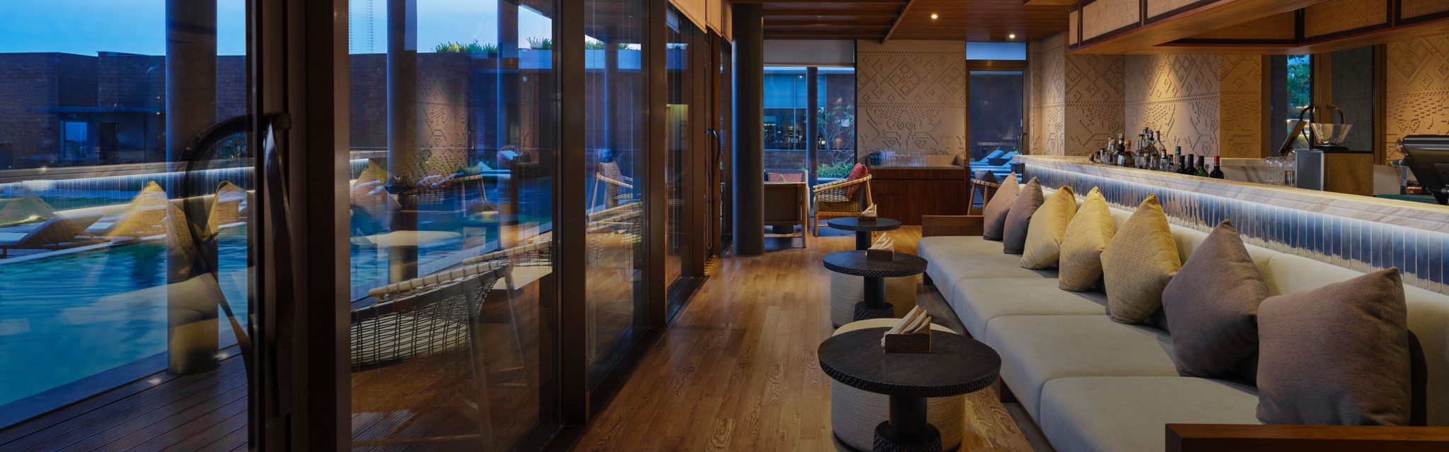 Luxurious indoor lounge with plush sofas and pillows beside a large window overlooking a pool. Warm lighting creates a cozy ambiance.
