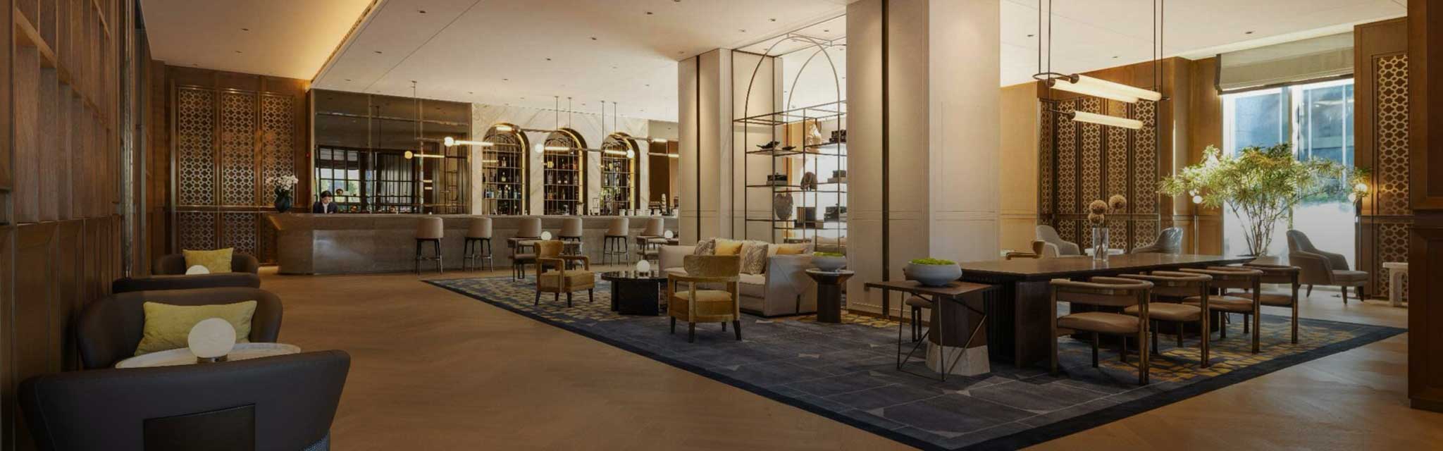 Spacious, elegant hotel lounge with modern decor. Features seating areas, wooden accents, a long bar, large windows, and warm lighting, creating a welcoming ambiance.