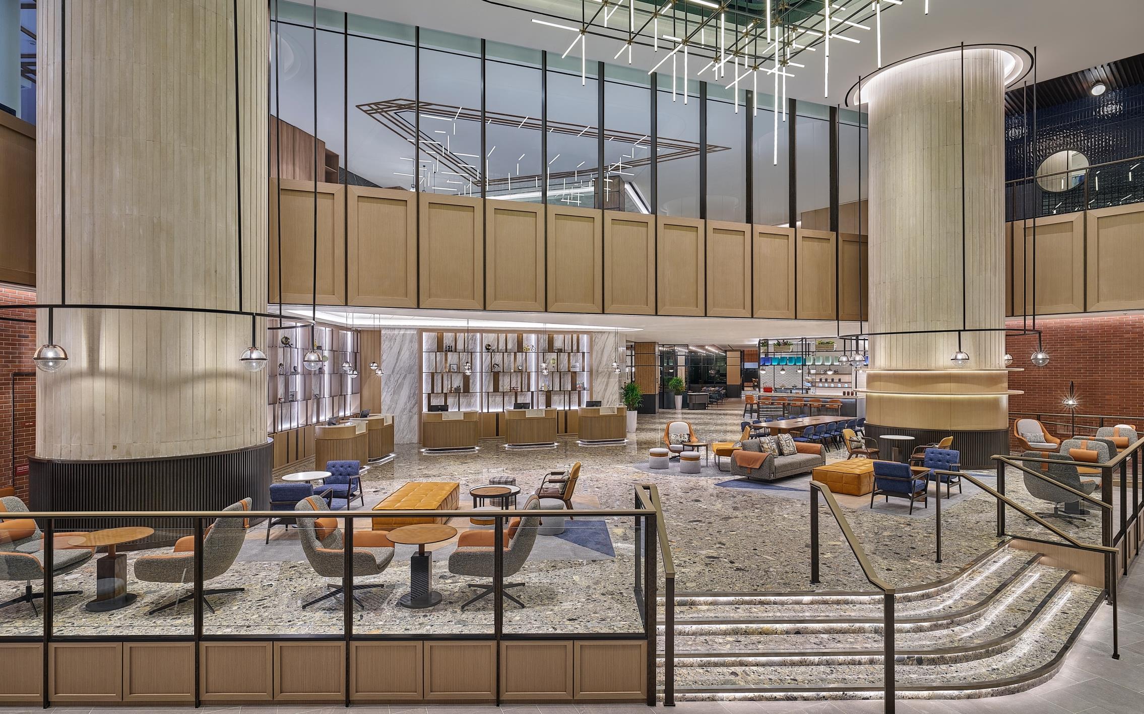 Luxurious hotel lobby with modern design, featuring tall columns, a glass and metal staircase, sleek furniture, and ambient lighting. Warm and inviting tone.
