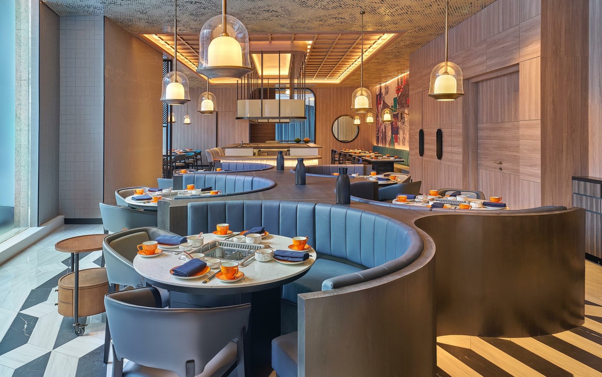 A modern restaurant interior with curved blue booths and round tables set with orange cups and blue napkins. Warm pendant lights create a cozy atmosphere.