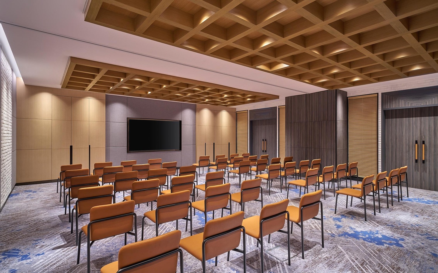 Modern conference room with rows of orange chairs facing a large flat-screen TV on a wooden panel wall. Ceiling has a wooden grid design.
