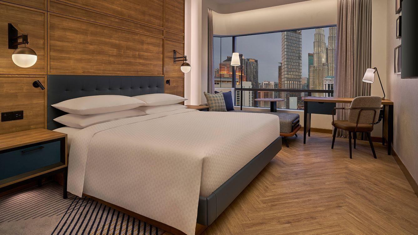 Modern hotel room with a cozy king-sized bed, stylish wood paneling, and large window revealing a cityscape. Warm lighting creates a welcoming ambiance.