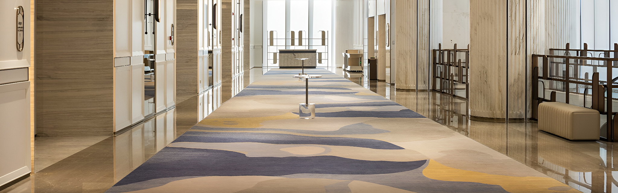 Wide hotel corridor with elegant abstract carpet, mirrored panel walls, and soft lighting. The atmosphere is serene and spacious, leading to large windows.