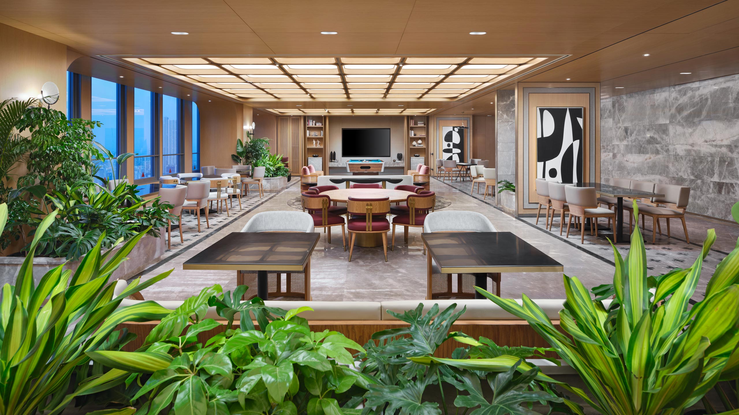 A modern, spacious lounge with a warm wood ceiling, geometric lighting, vibrant plants, and abstract art. It features cozy seating and large windows, creating an inviting ambiance.