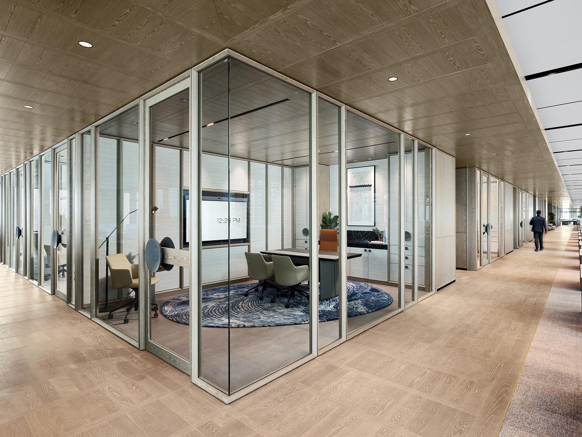 Stylish modern office with glass walls, minimalist furniture, and a blue-patterned rug. A lone person walks down the hallway, conveying a calm, professional atmosphere.