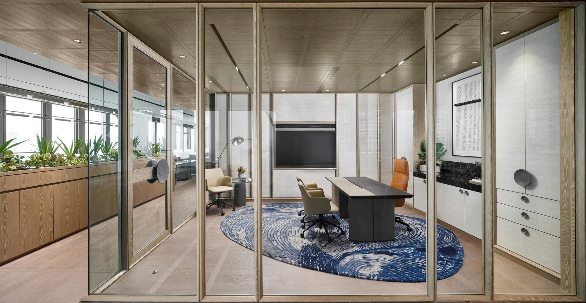 Modern office with glass walls, wooden accents, and a large desk on a blue wave-patterned rug. Neutral tones and plants create a calm, professional atmosphere.