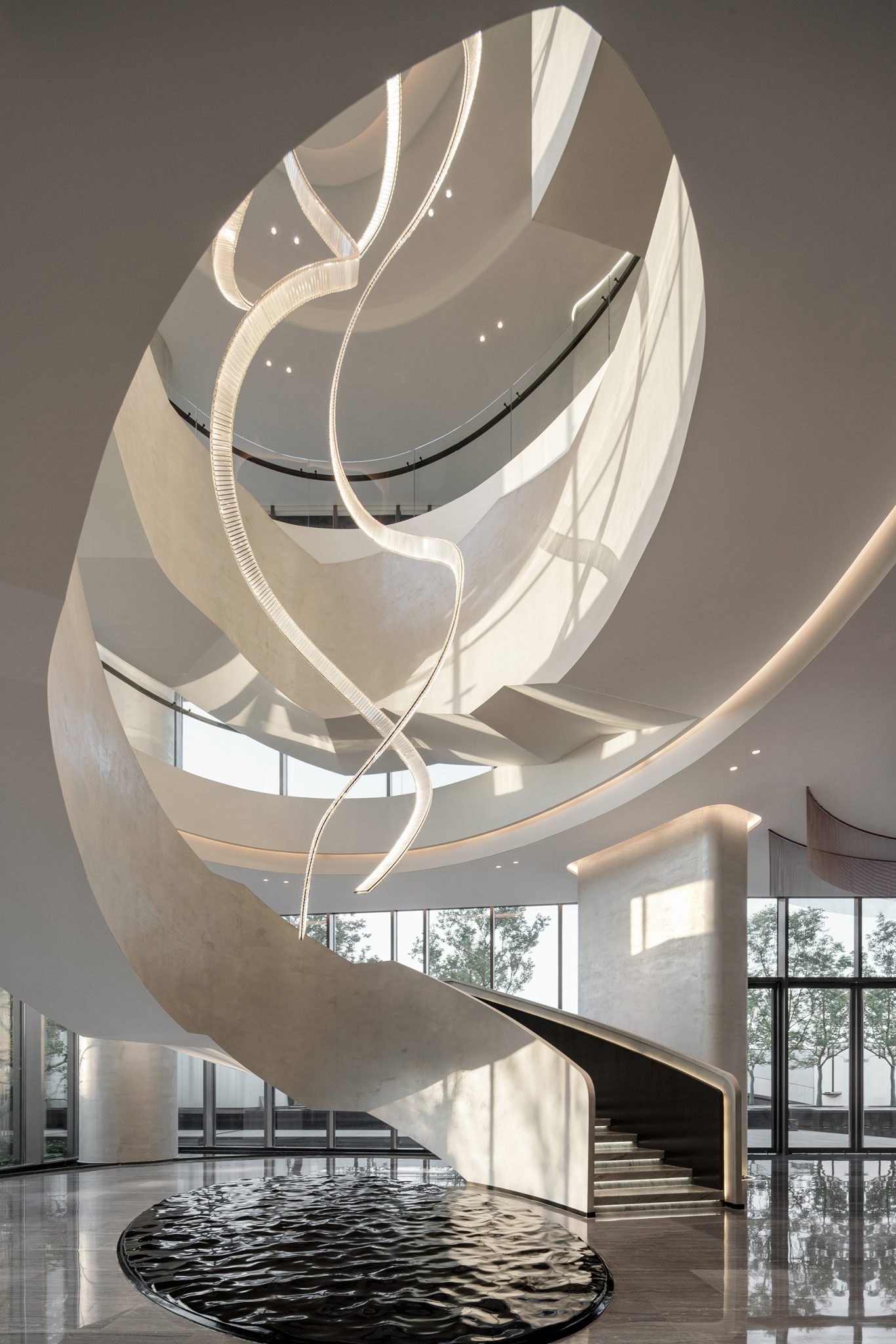 A grand, elegant spiral staircase with minimalist design and smooth curves centers a modern interior. A large window reveals trees outside. Light streams in, creating soft shadows.