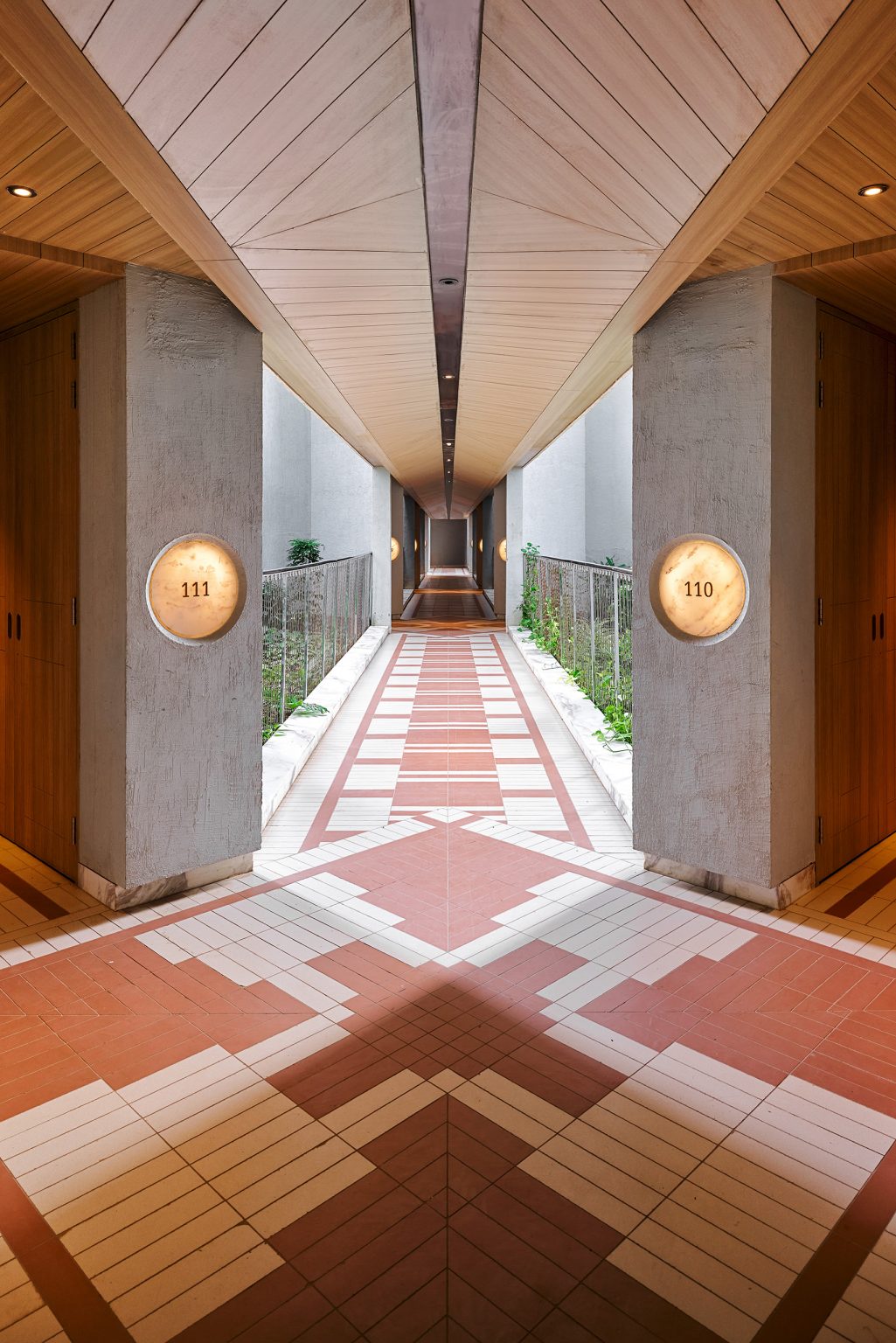 A symmetrical corridor with geometric red and tan tiled flooring leads into the distance. Concrete pillars flank each side with round wall lights numbered 111 and 110, creating a modern and serene atmosphere.