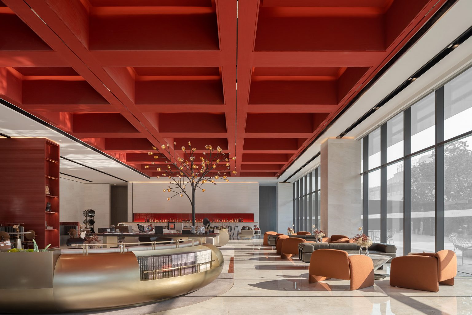 Spacious modern lobby with a bold red ceiling, large windows, and contemporary seating. A decorative tree centerpiece adds elegance and warmth.