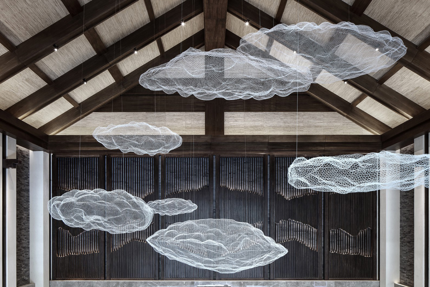 Suspended wire mesh clouds float under a high, wooden, loft-style ceiling in a dimly lit, modern interior, creating a serene and airy atmosphere.