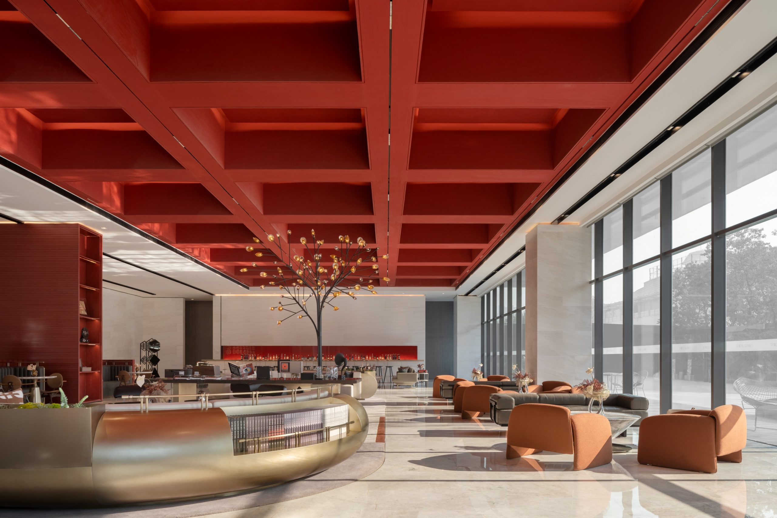 Spacious modern lobby with high red coffered ceiling, large windows, and a central tree sculpture. Brown chairs and golden accents create a warm, elegant atmosphere.