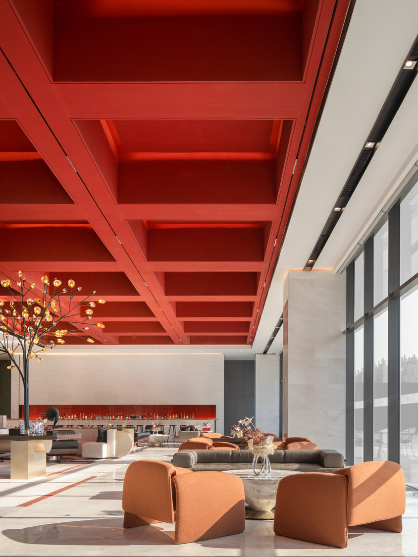 Spacious modern lobby with a striking red coffered ceiling, orange seating, large floor-to-ceiling windows, and a decorative tree with lights.