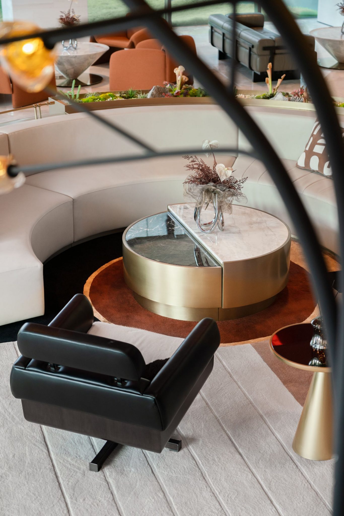 A modern lounge area featuring a circular coffee table with a glass top and a gold base, surrounded by a white curved seating arrangement. A black chair is positioned nearby, and there are decorative elements like flowers on the table.