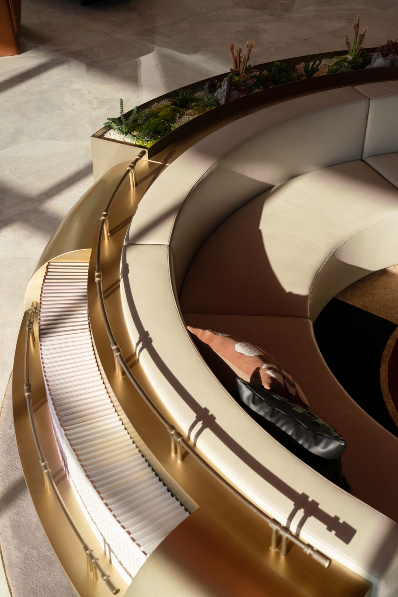 A circular, elegantly designed seating area featuring light-colored upholstery with gold accents, surrounded by greenery.