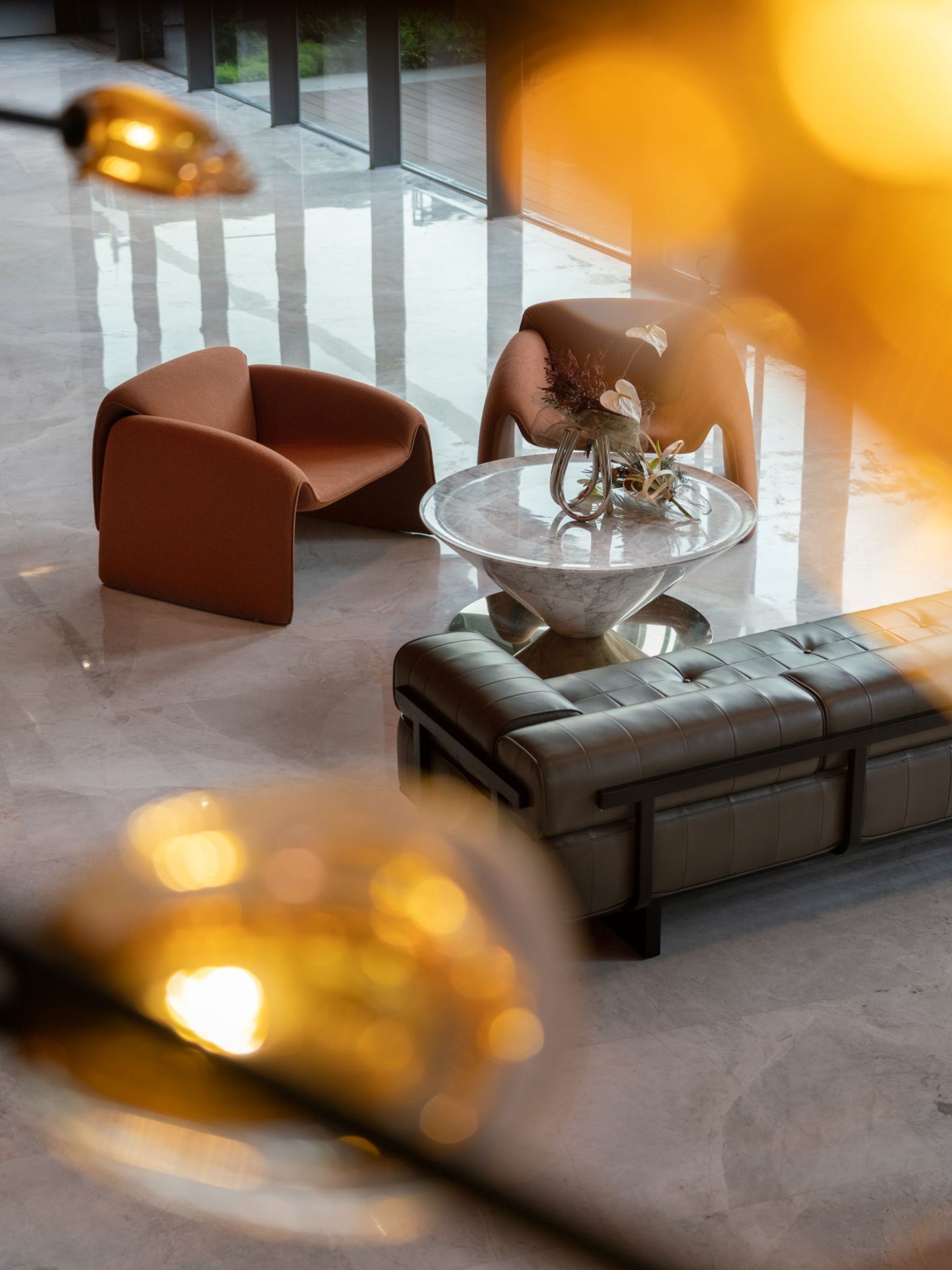 A modern interior space with two orange chairs, a glass coffee table with a decorative bowl, and a dark leather sofa, softly illuminated by overhead lights.