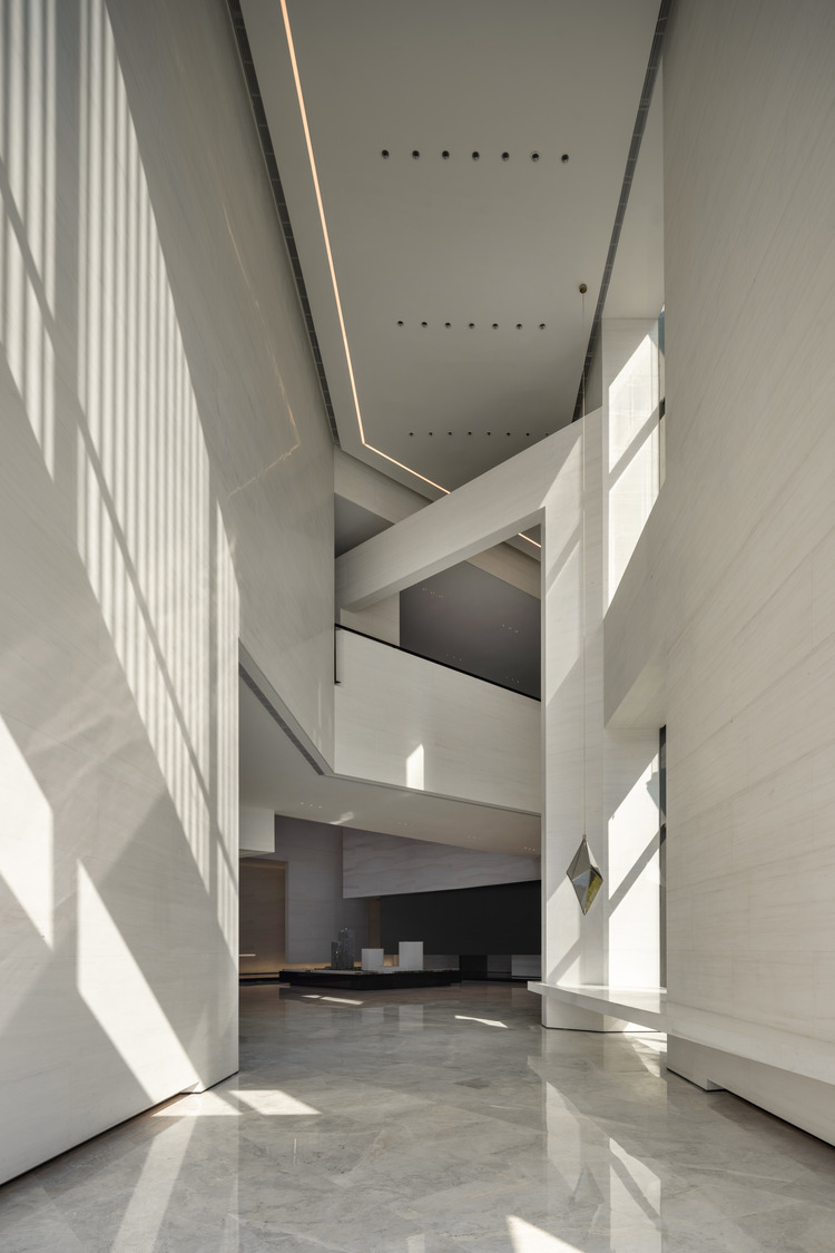A modern architectural interior with white walls, angular lines, and ample natural light creating shadows on the marble floor.