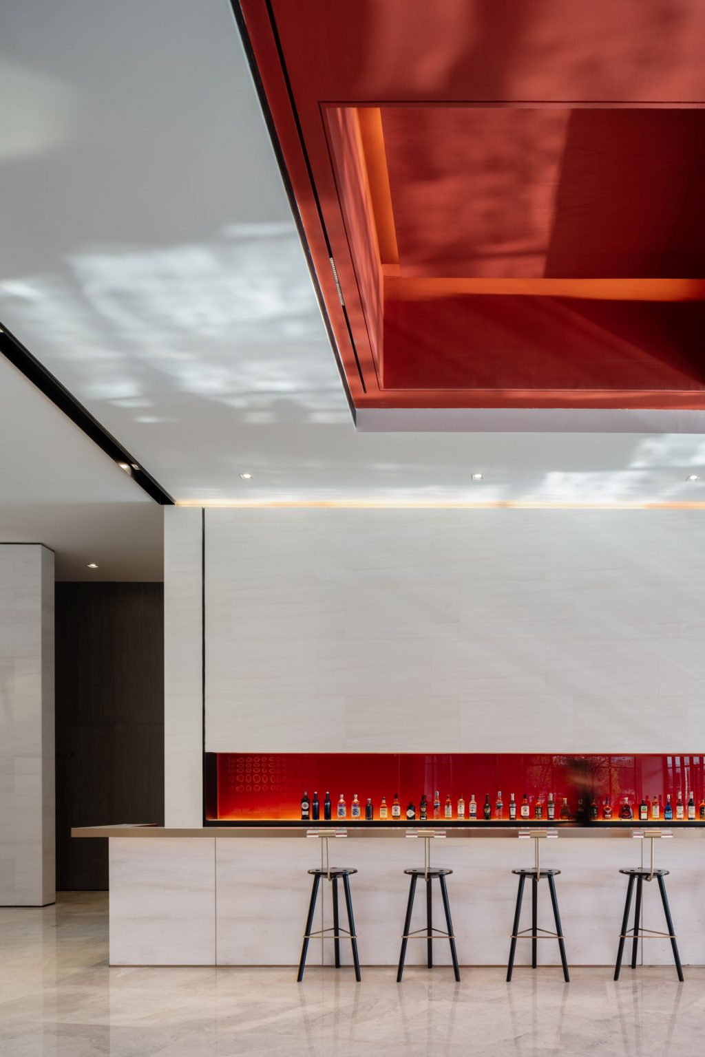 A modern bar area with a minimalist design, featuring a long counter, high stools, and a backlit shelf displaying various bottles, set against a sleek white wall and a bold red ceiling accent.
