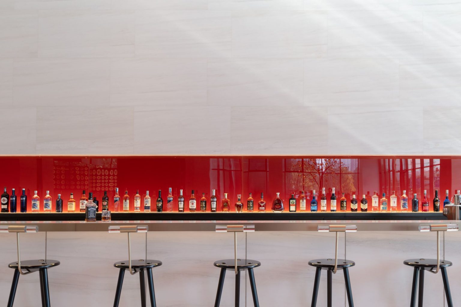 A sleek bar with a row of various liquor bottles displayed against a red backdrop, accompanied by tall stools.