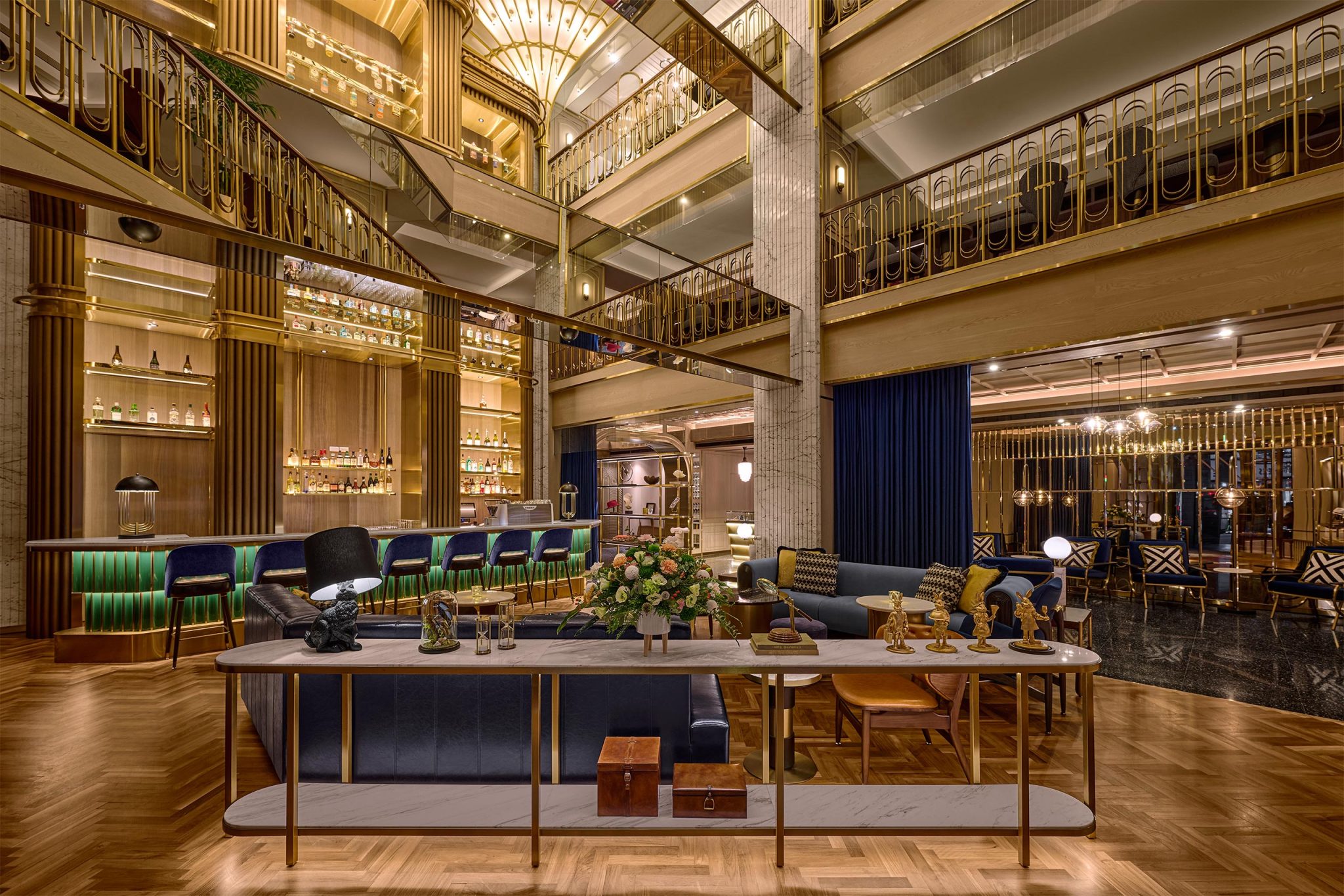 Luxurious Art Deco hotel lobby with a glowing bar, gold railings, velvet seating, floral tables, and warm, inviting lighting.
