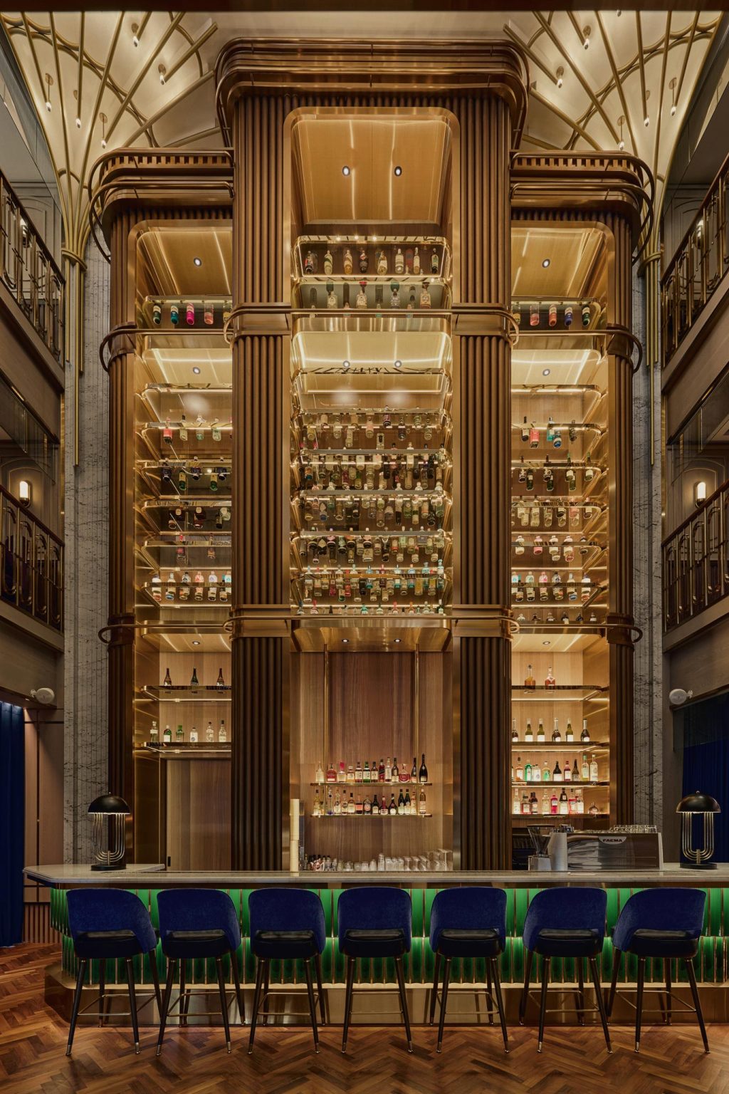 Symmetrical art deco cocktail bar with glowing shelves of liquor bottles, ornate woodwork, green velvet stools, and a warm, luxurious atmosphere.