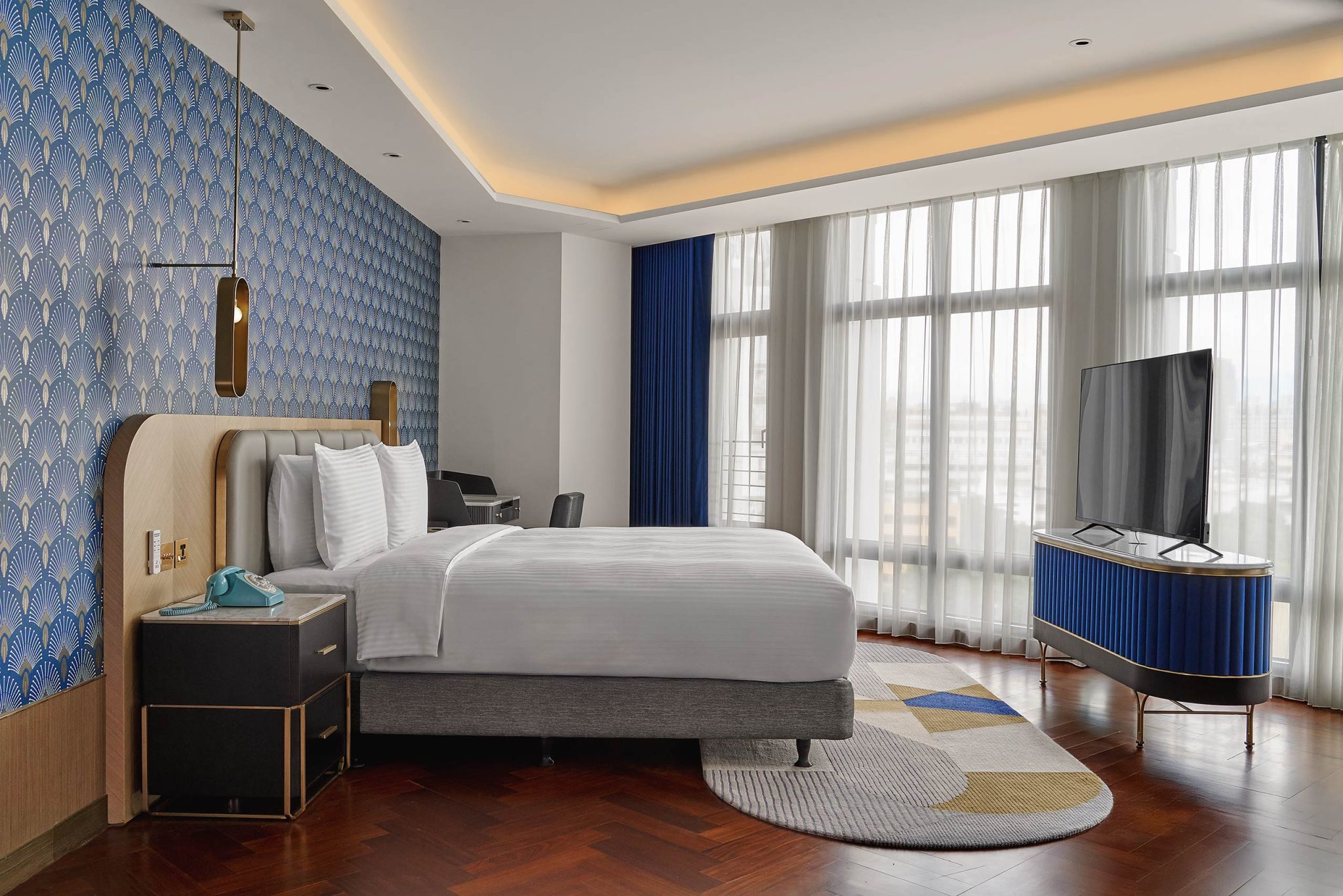 Modern hotel bedroom with patterned blue wallpaper, plush bed, floor-to-ceiling windows, TV console, and warm, elegant decor.