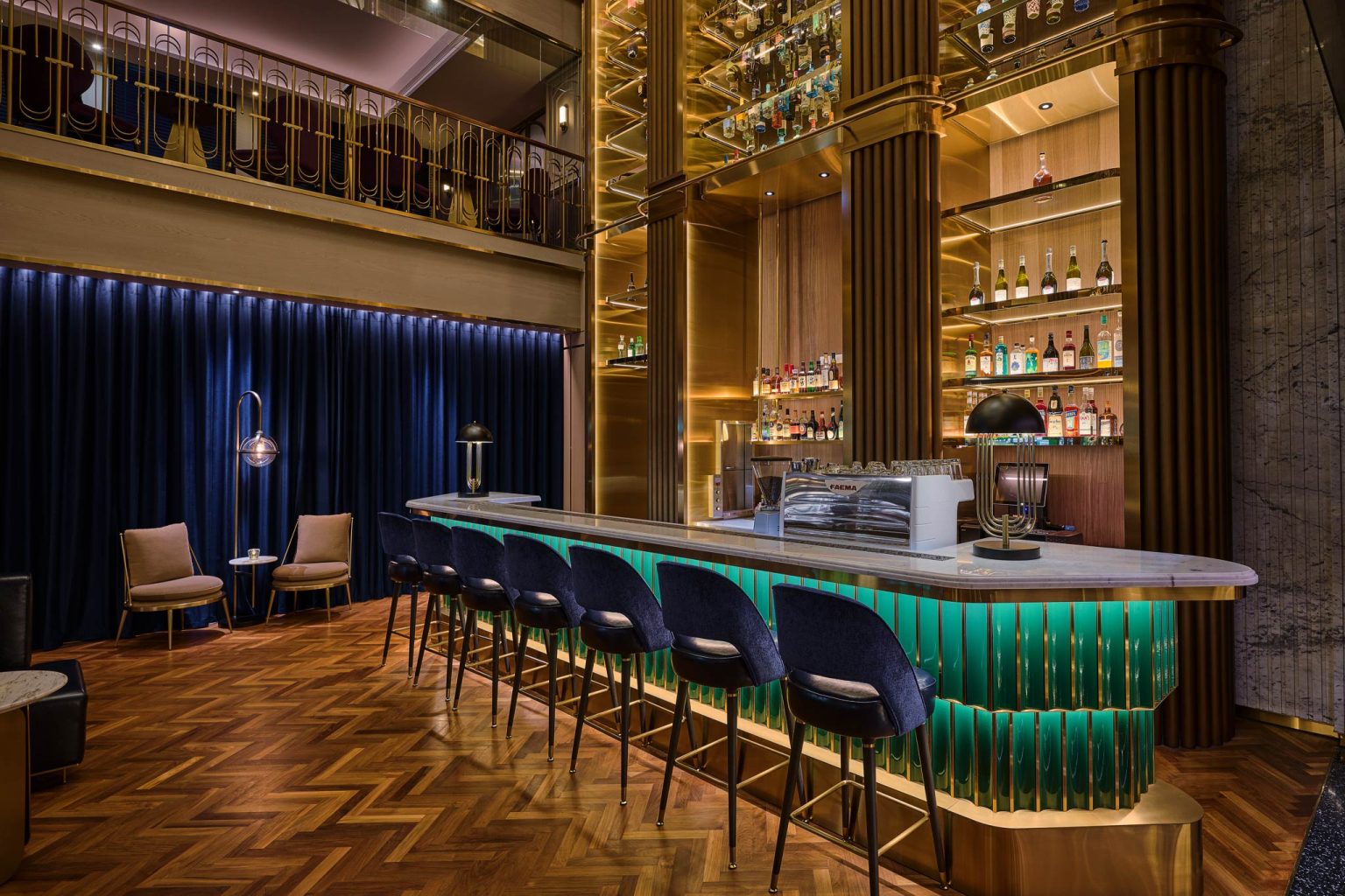 Luxurious cocktail bar with emerald-green counter, navy velvet seating, warm brass accents, glowing shelves, and herringbone wood floors.